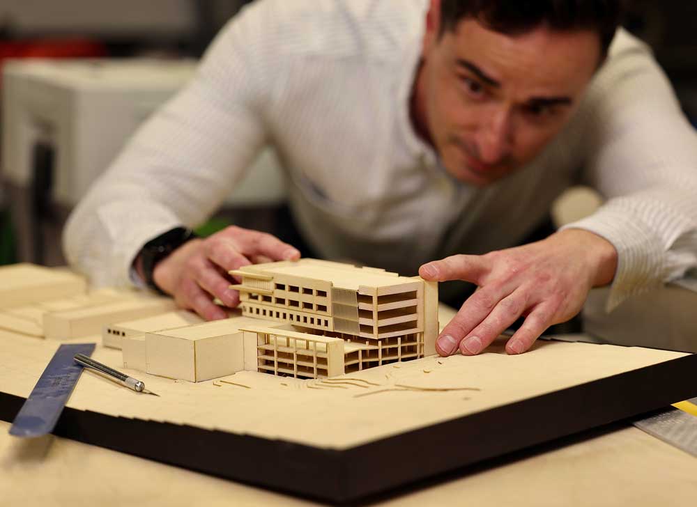 An MOA architect leans in closely to examine a detailed balsa wood architectural scale model of a multi-story building, with drafting tools visible on the table beside it.