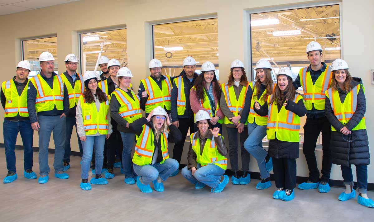 The MOA Architecture team posing cheerfully on a construction site walkthrough, wearing white hard hats, yellow safety vests, and blue protective shoe covers i…The MOA Architecture team posing cheerfully on a construction site walkthrough, wearing white hard hats, yellow safety vests, and blue protective shoe covers inside a nearly completed building.