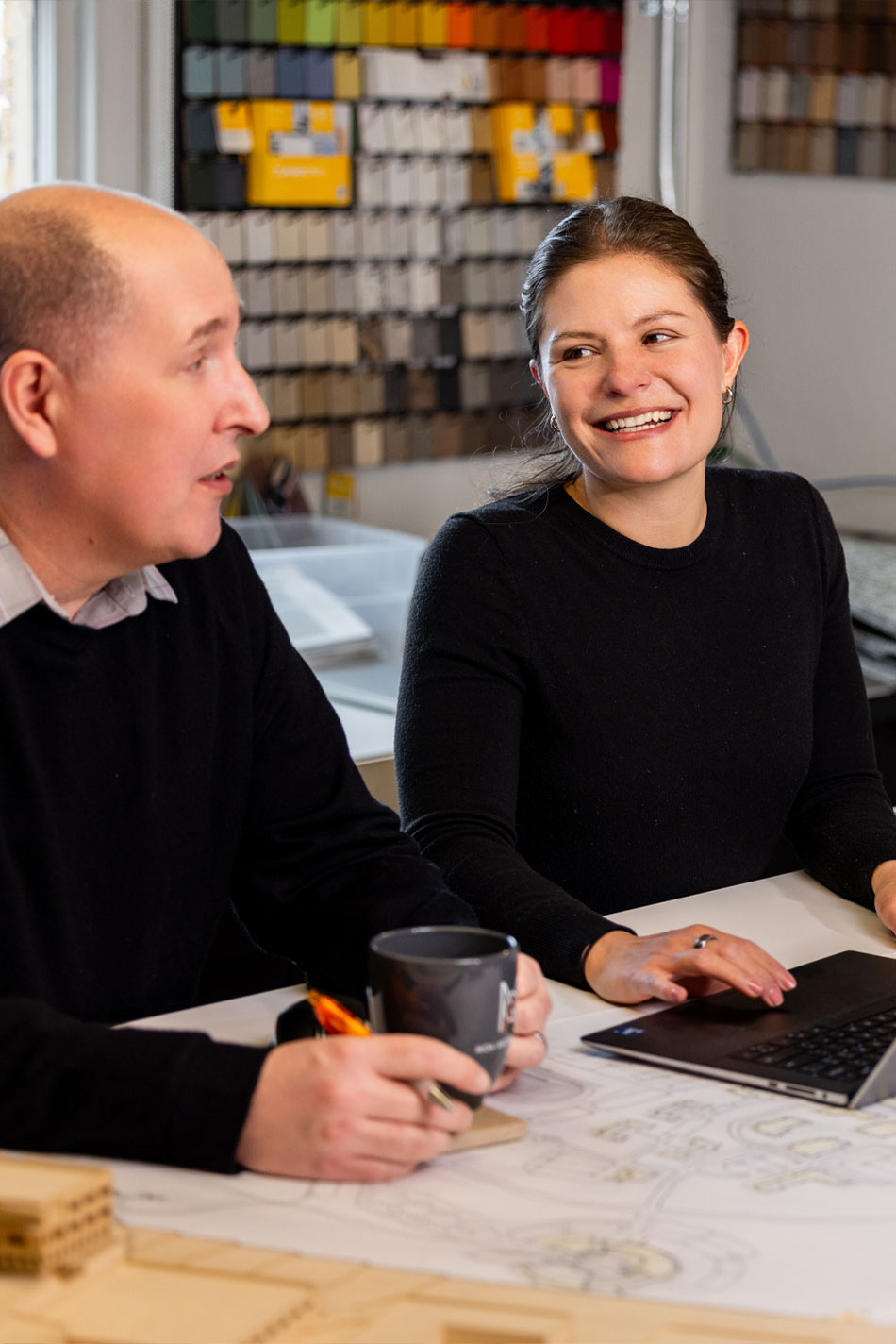 Two MOA team members collaborating at a design studio table with a wall of material samples behind them.