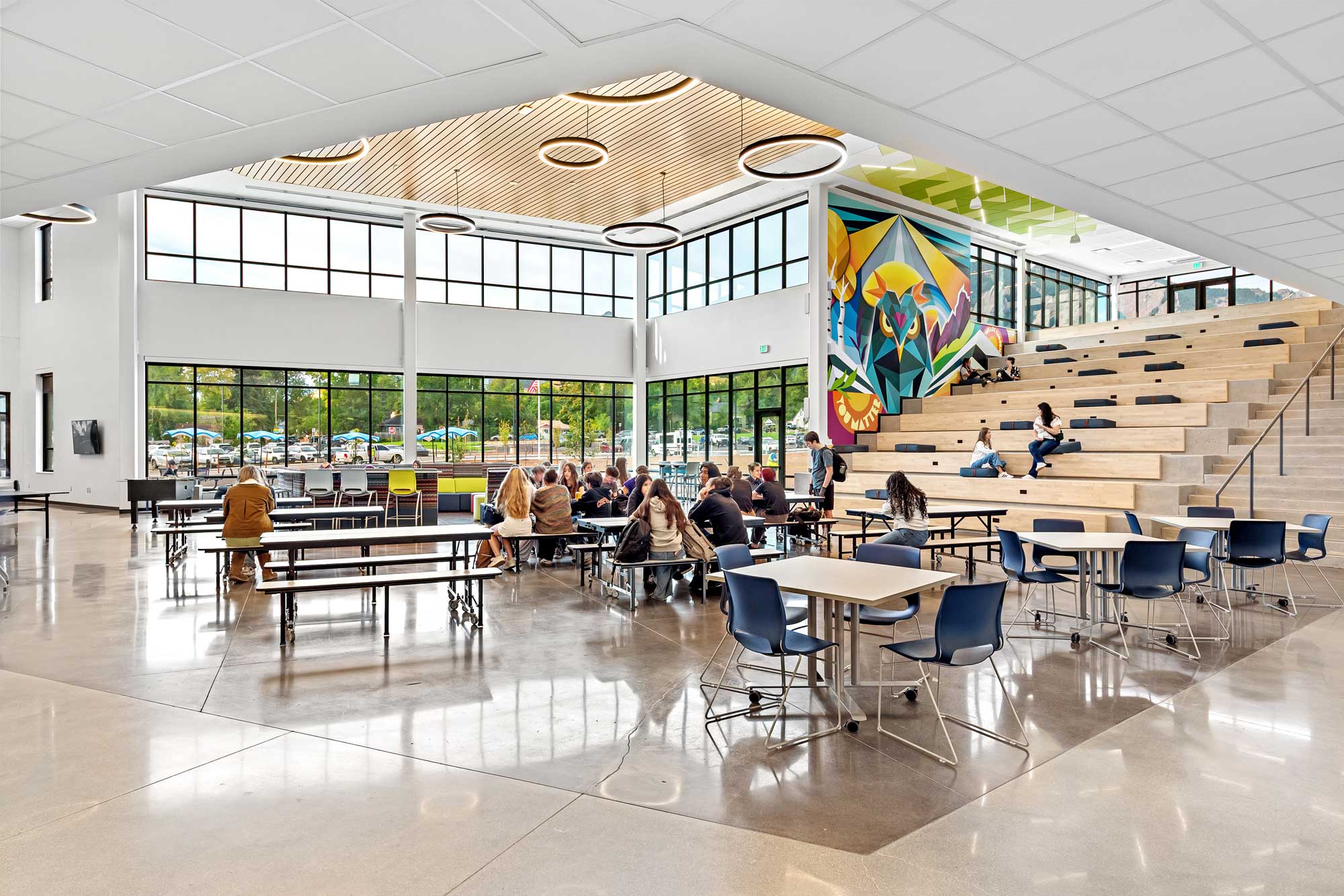 Modern school commons with learning stairs, a geometric owl mural, and floor-to-ceiling windows.