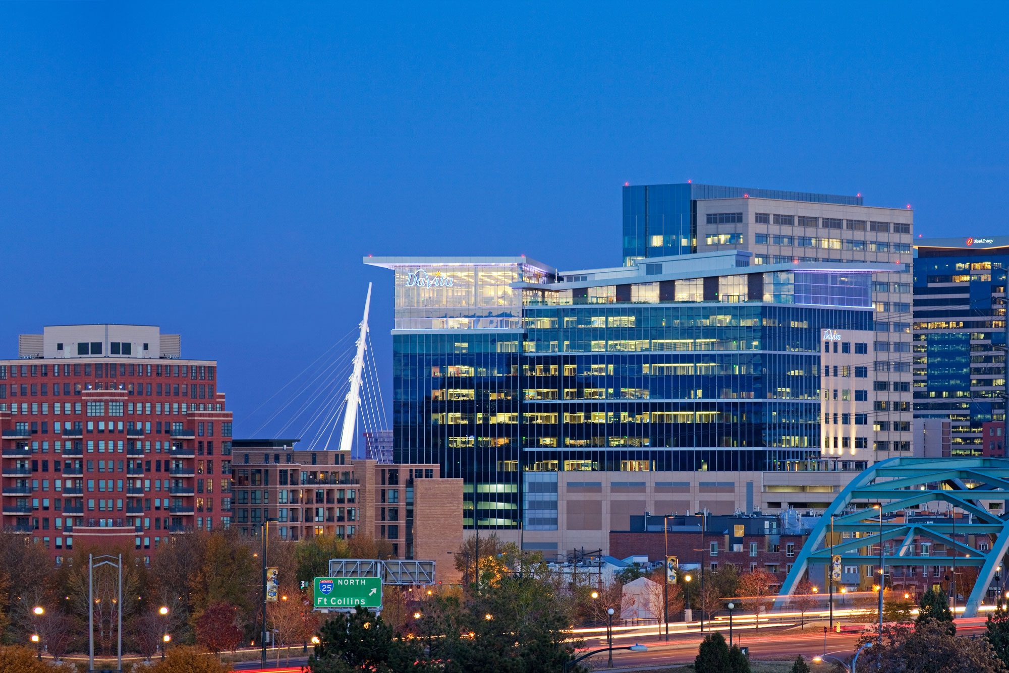 The DaVita headquarters building illuminated at dusk against the Denver skyline, with a glass-topped penthouse level, a cable-stayed bridge, and light trails from highway traffic in the foreground.