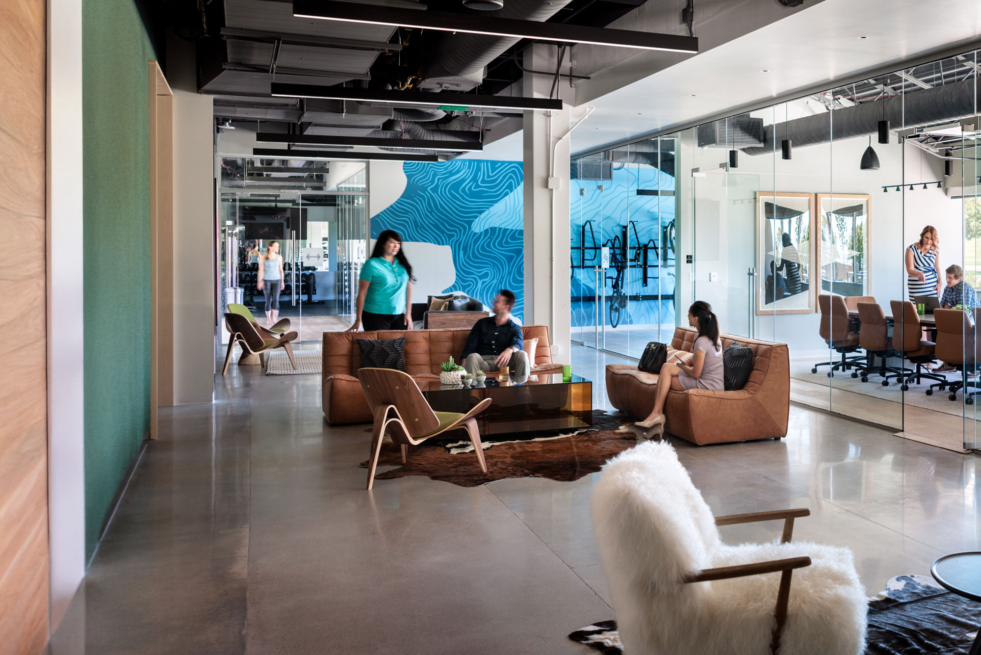 A modern office lounge at Infinite Labs with people gathered on leather sofas and designer chairs, glass-walled conference rooms, an exposed black ceiling, and…A modern office lounge at Infinite Labs with people gathered on leather sofas and designer chairs, glass-walled conference rooms, an exposed black ceiling, and a bold blue topographic mural in the background.